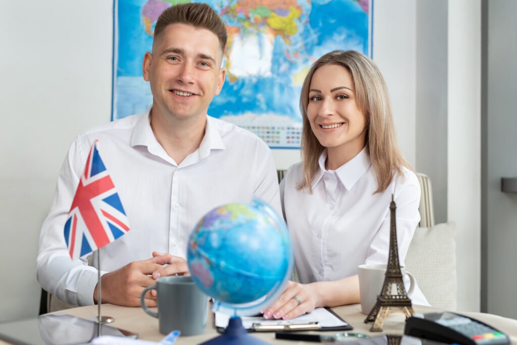 Zwei junge Menschen sitzen lächelnd an einem Tisch mit Weltkarte, Globus und UK-Flagge – ein motivierendes Umfeld, um effizient Englisch lernen zu können.